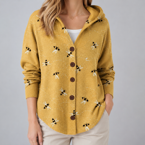A woman wearing a mustard yellow hooded cardigan with a bumblebee pattern and brown buttons, paired with a white top and light beige pants, against a solid gray background.