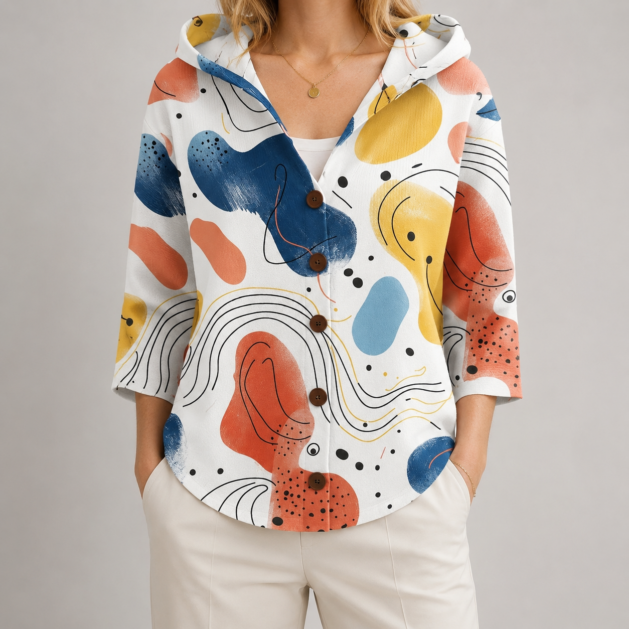 A woman wearing a white hooded button-front top with a colorful abstract print in blue, yellow, orange, and coral, paired with light beige pants, against a plain light gray studio background.