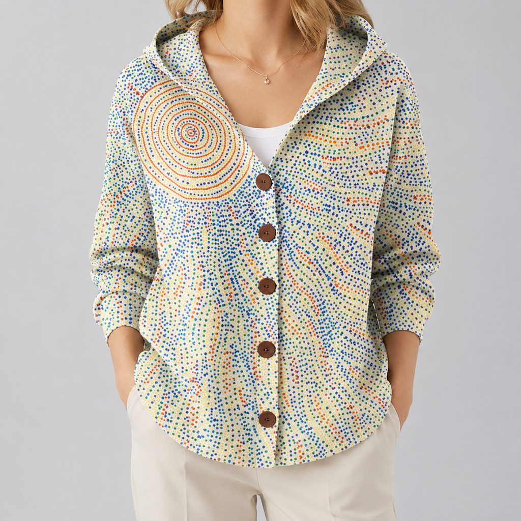 A woman wearing a hooded button-front cardigan with a multicolor dotted pattern and a large circular spiral motif on the left chest, paired with light beige pants against a plain light gray studio background.