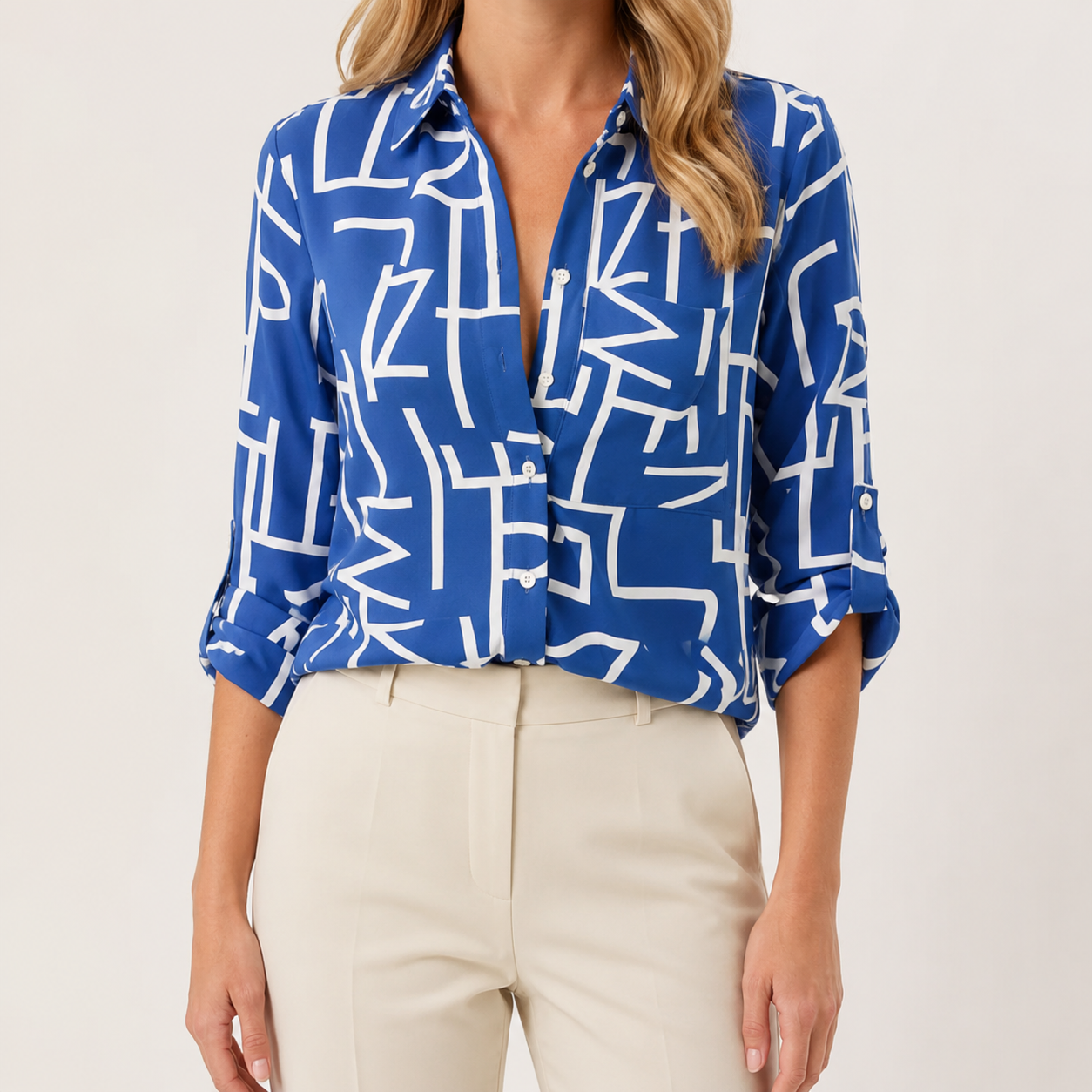 A woman wearing a blue button-up blouse with a white abstract geometric line pattern, paired with light beige trousers, shown against a plain off-white studio background.