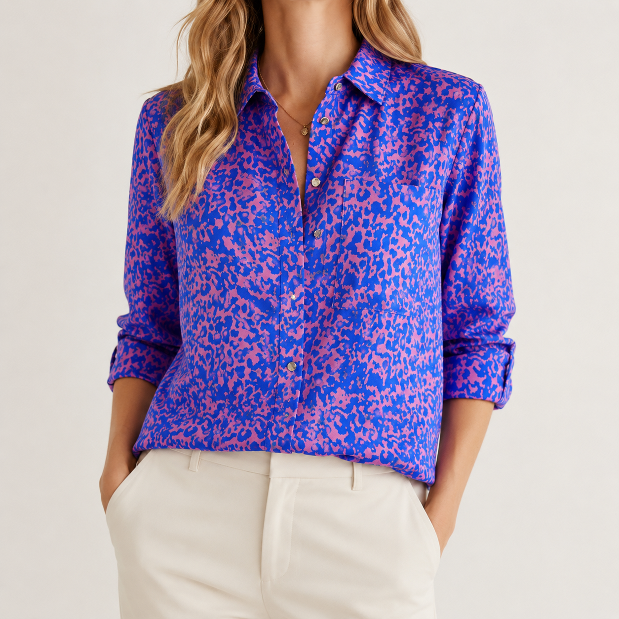 A woman wearing a long-sleeve button-up shirt with a blue and pink abstract animal print, paired with off-white trousers, against a plain light background.