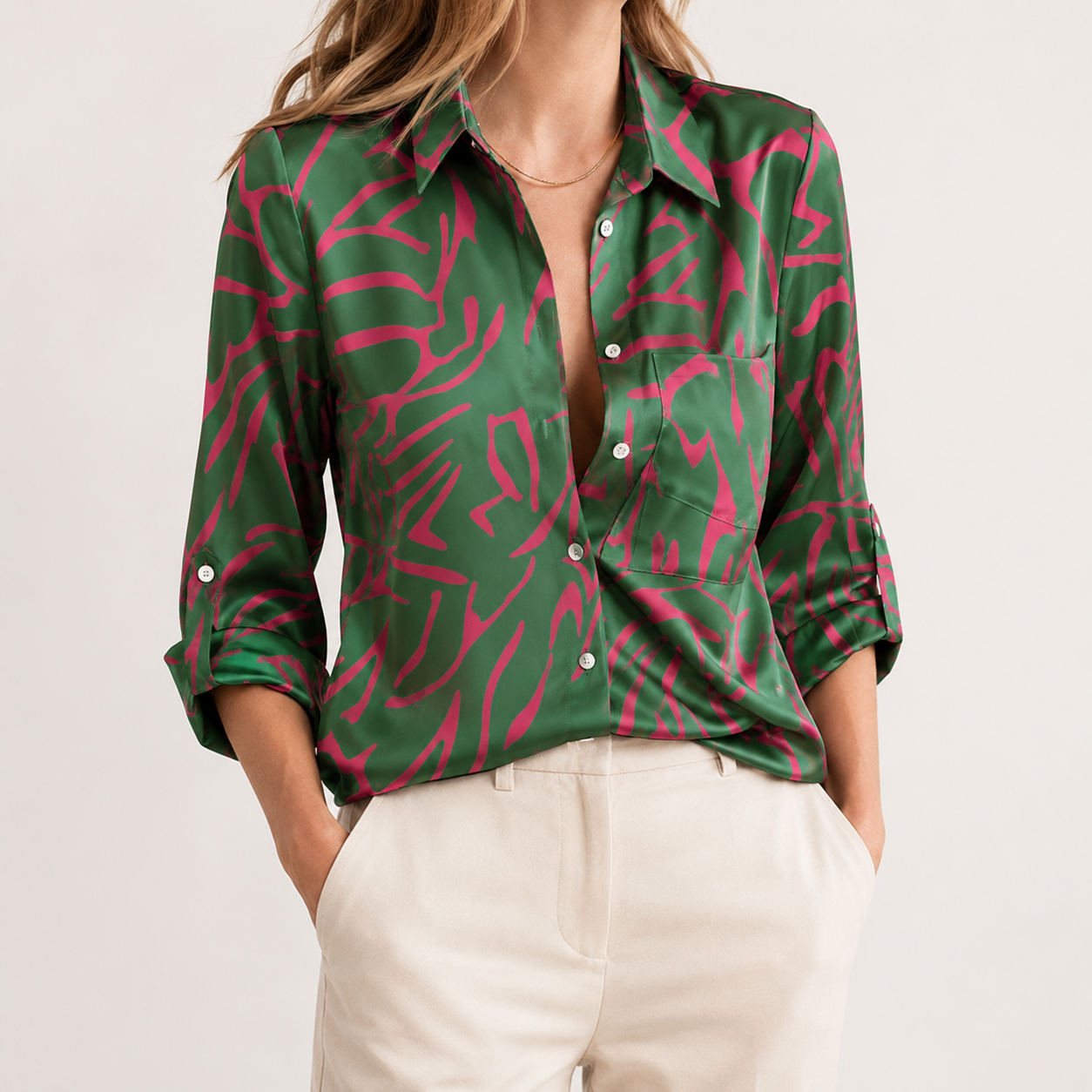 A woman wearing a green satin button-up shirt with a vibrant pink abstract pattern, paired with cream-colored trousers, against a plain light background.