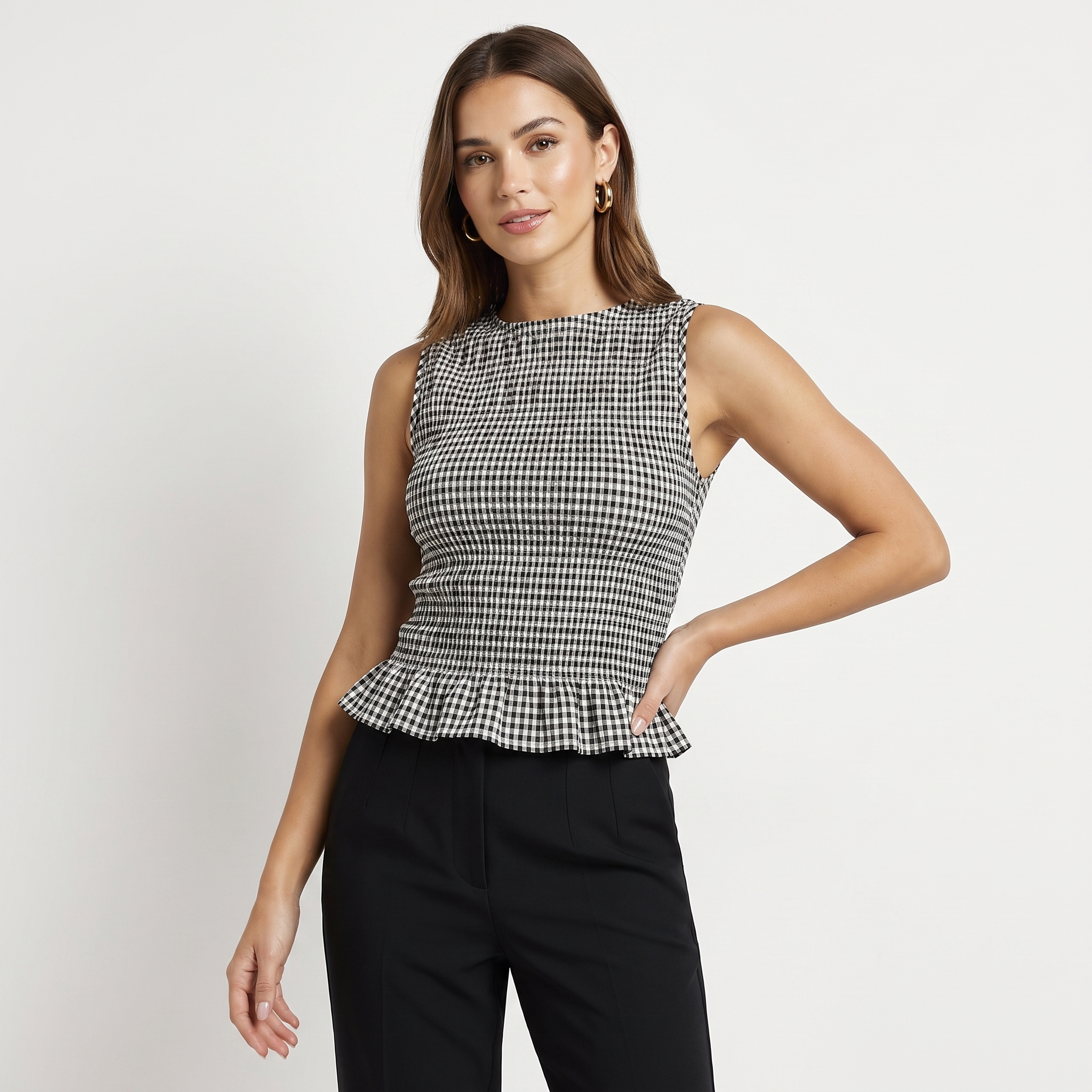 A woman modeling a sleeveless black and white gingham peplum top paired with black trousers, photographed against a clean light gray studio background.