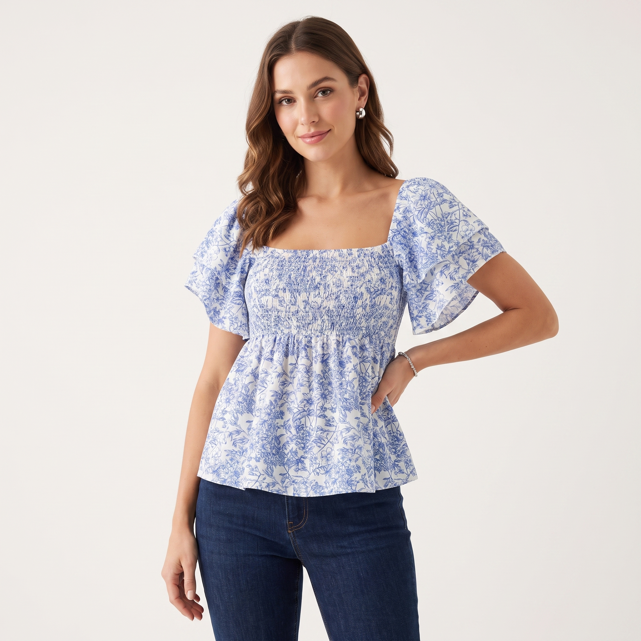 A woman modeling a blue and white floral print smocked blouse with puff sleeves and a peplum hem, paired with dark blue jeans, against a plain off-white studio background.