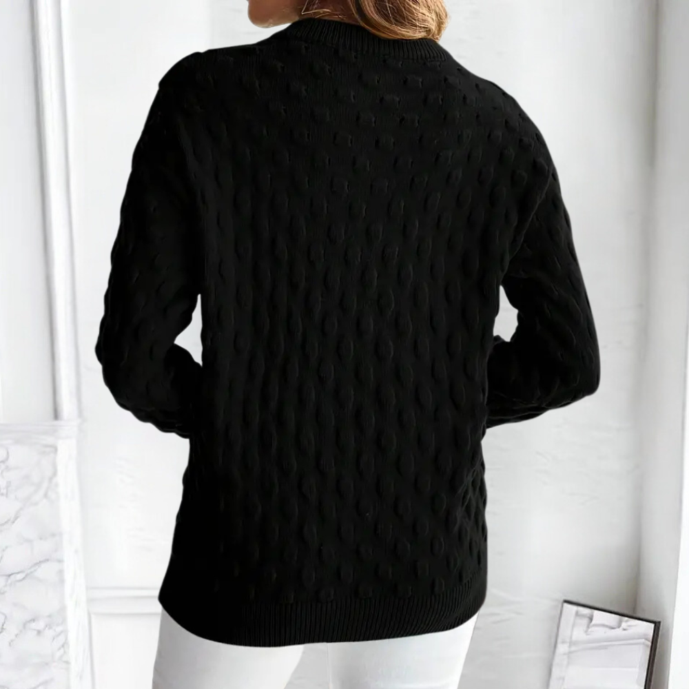 Back view of a black cable knit crew neck sweater with long sleeves and ribbed cuffs, worn over white trousers.