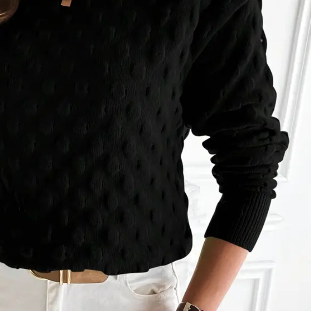 Black cable knit crew neck sweater with long sleeves and ribbed cuffs, styled with white trousers.