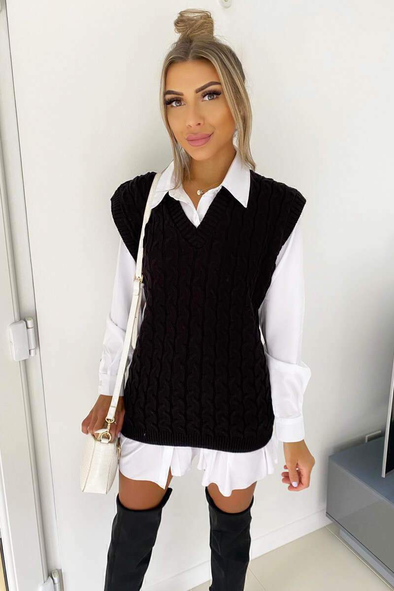 Woman wearing a black cable knit V-neck sweater vest layered over a crisp white collared shirt and black over-knee boots.