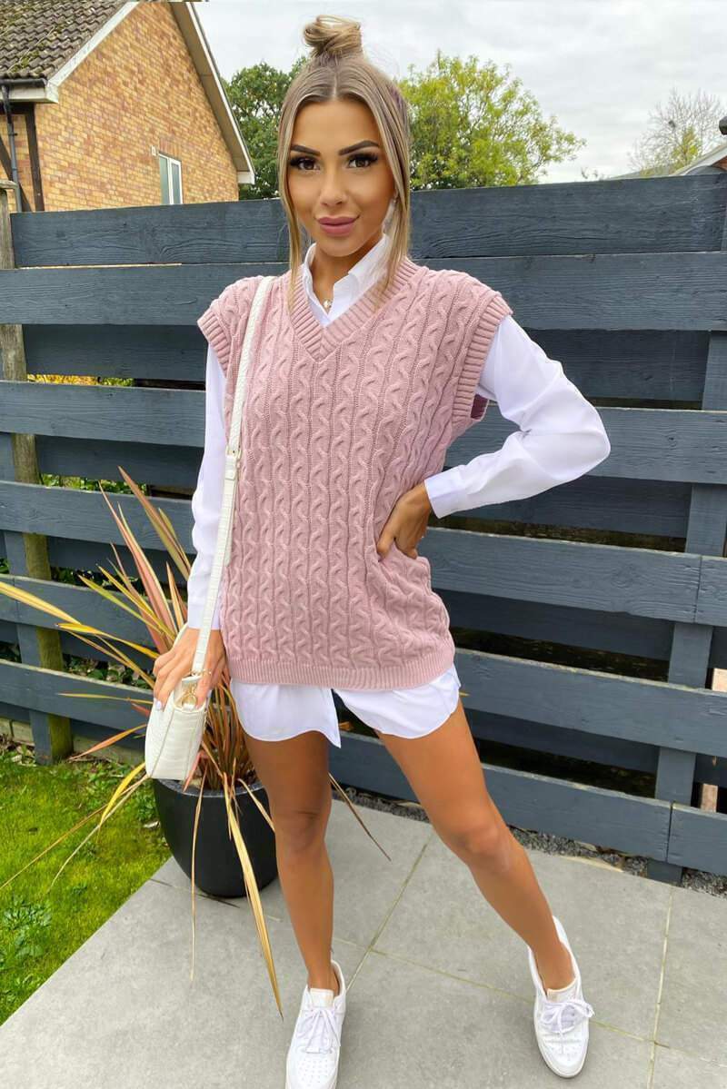 Woman wearing a dusty pink cable knit V-neck vest layered over a crisp white button-down shirt with white sneakers.