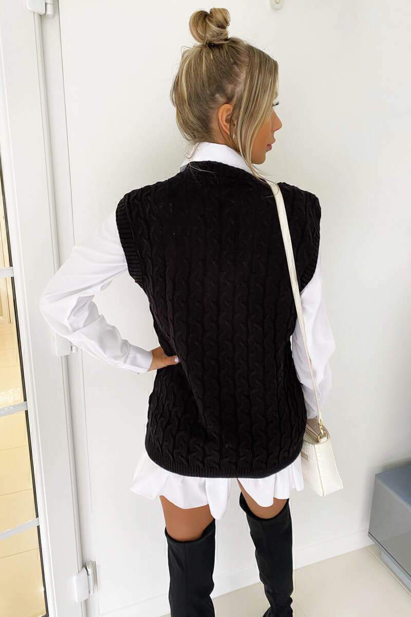 Woman wearing a black cable knit vest layered over a crisp white shirt with black over-the-knee boots.