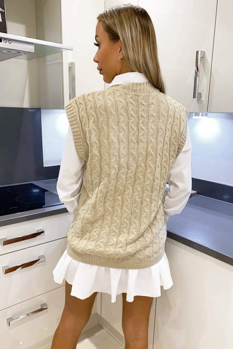 Beige cable knit sweater vest layered over a white collared shirt in a modern kitchen setting.
