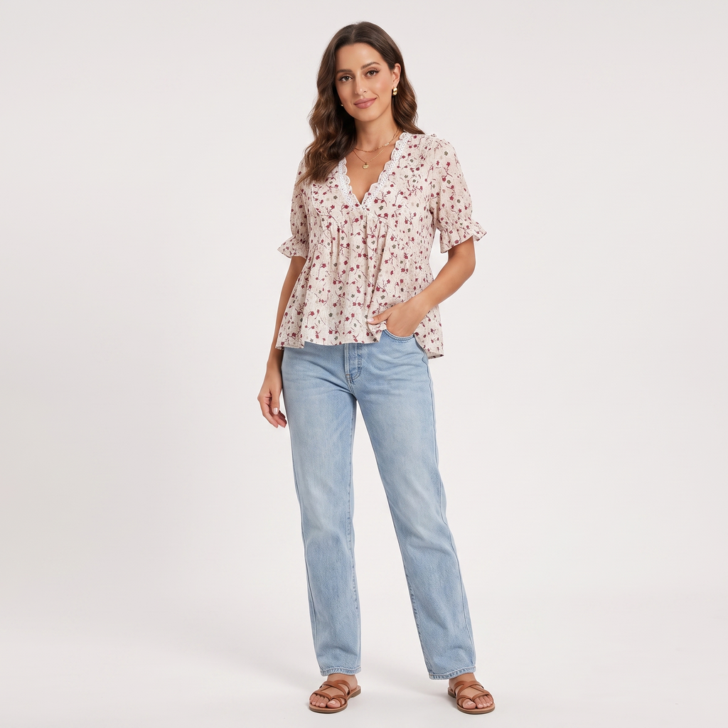 A woman modeling a short-sleeve V-neck blouse with a small floral print in pink and green on a light beige background, paired with light-wash blue jeans and brown sandals, against a plain white studio background.