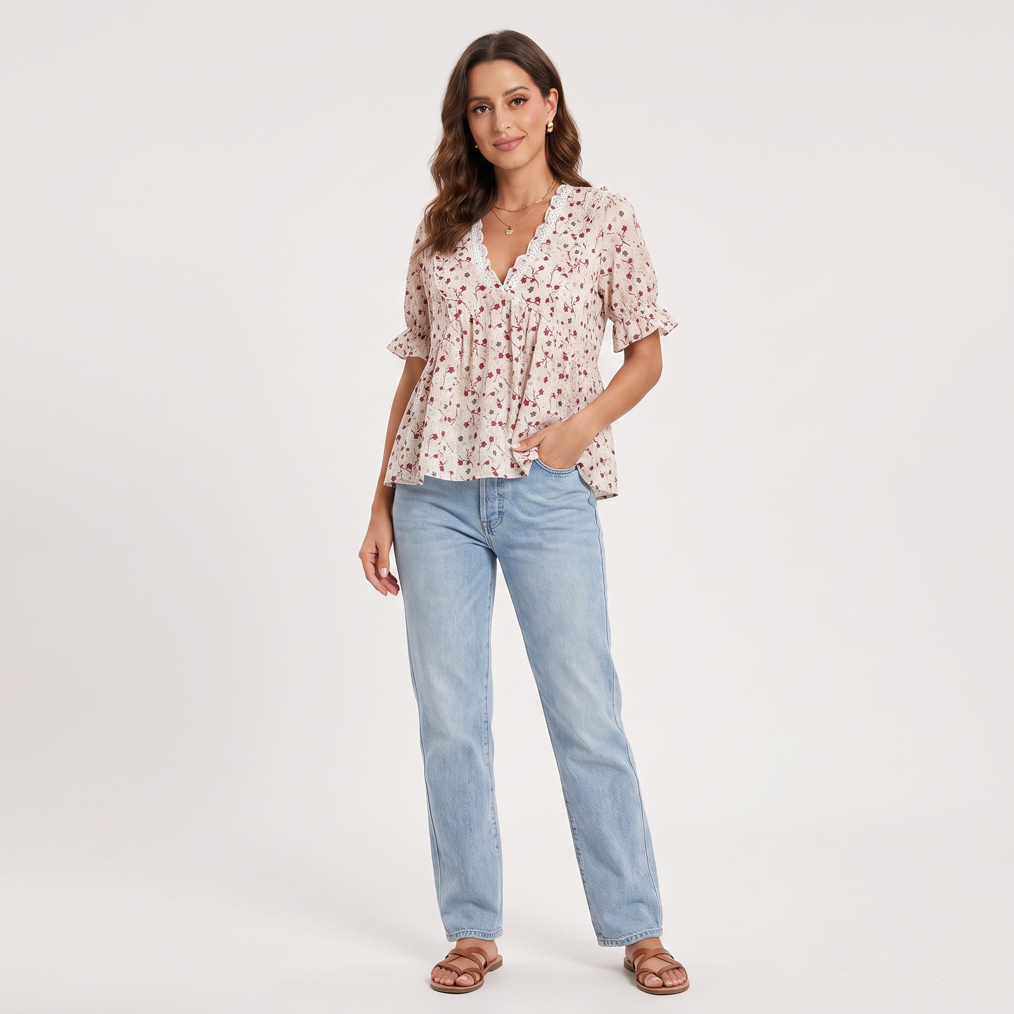 A woman modeling a short-sleeve V-neck blouse with a small floral print in pink and green on a light beige background, paired with light-wash blue jeans and brown sandals, against a plain white studio background.