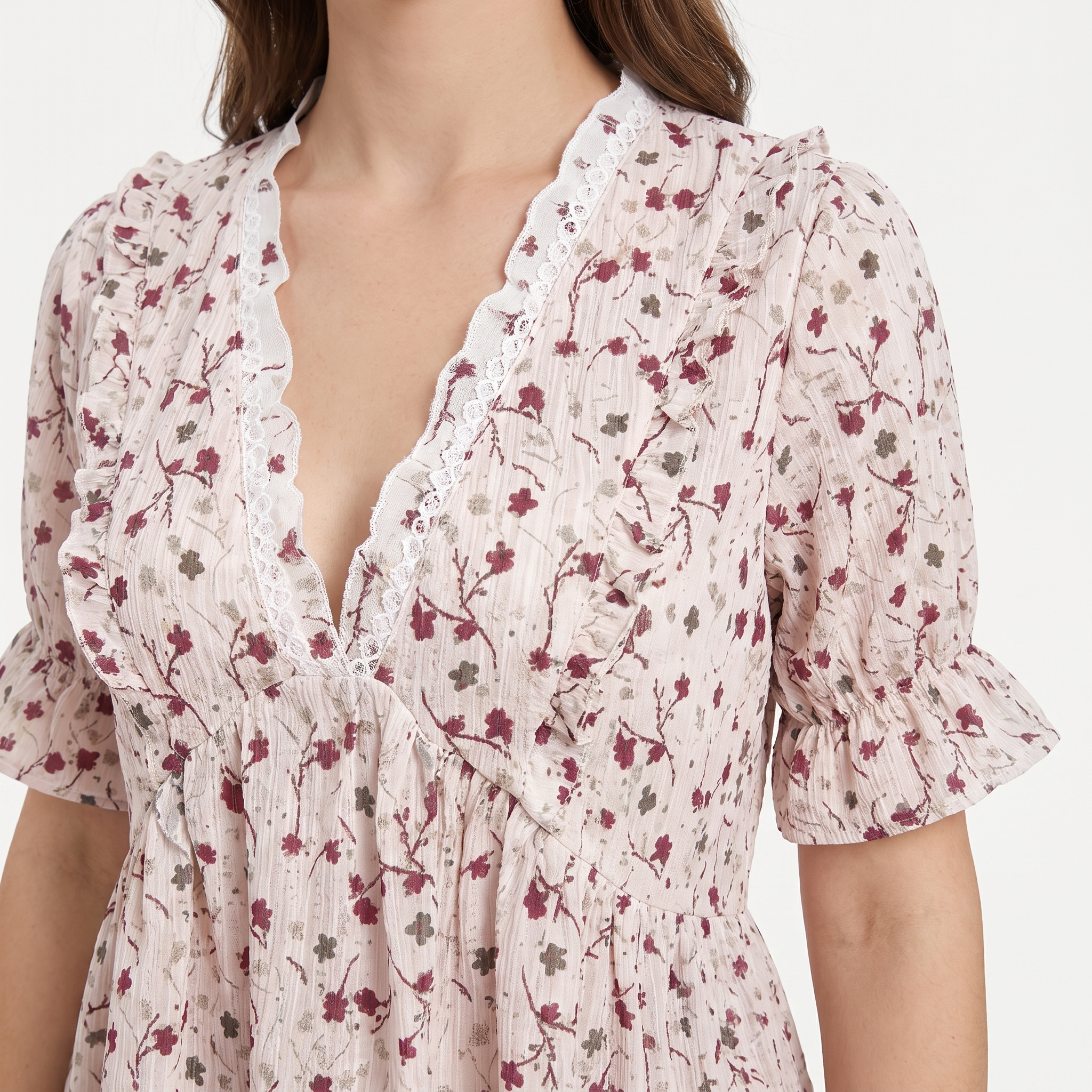 A close-up of a woman wearing a light pink crinkled dress with a dark red and olive floral print, featuring a ruffled lace-trimmed V-neckline and short puff sleeves.