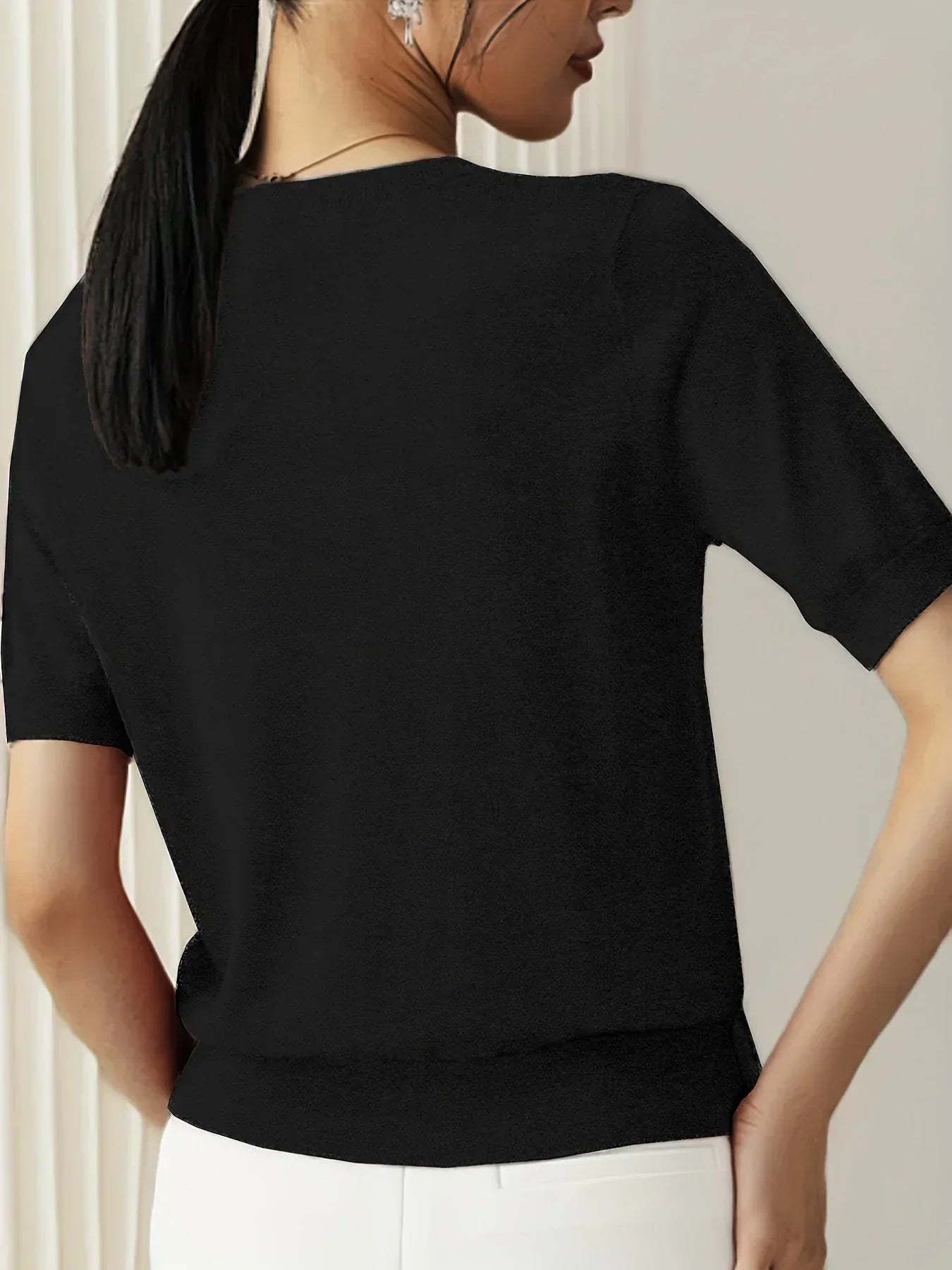 Black short-sleeve fine knit sweater with a crew neck and ribbed trim, worn with white trousers.