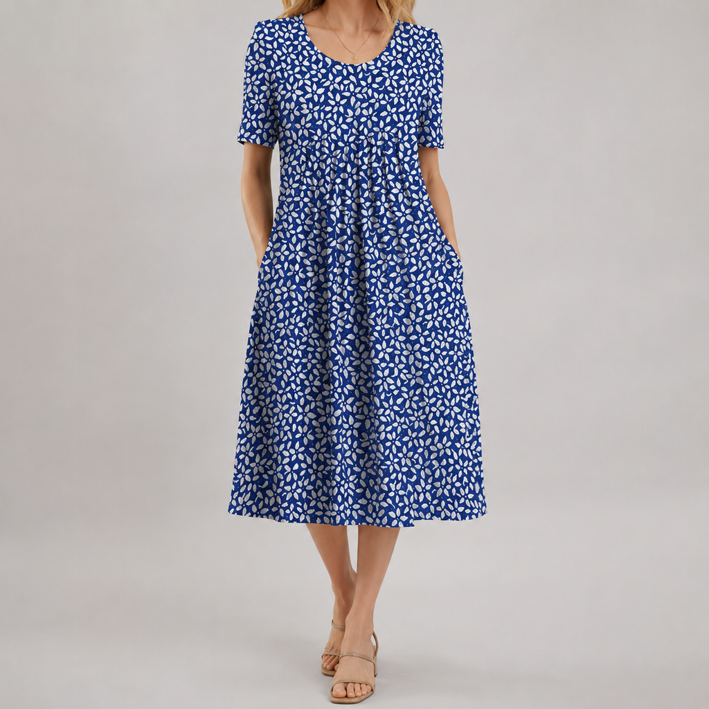 A woman wearing a short-sleeve midi dress with a blue and white floral pattern, side pockets, and a gathered waist, paired with beige flat sandals, photographed against a neutral gray studio background.