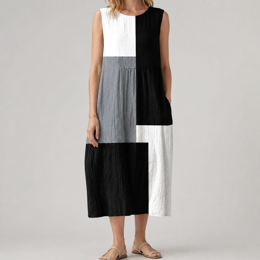 A woman wearing a sleeveless midi dress with a geometric patchwork design in black, white, and gray, paired with beige flat sandals, set against a plain light gray studio background.