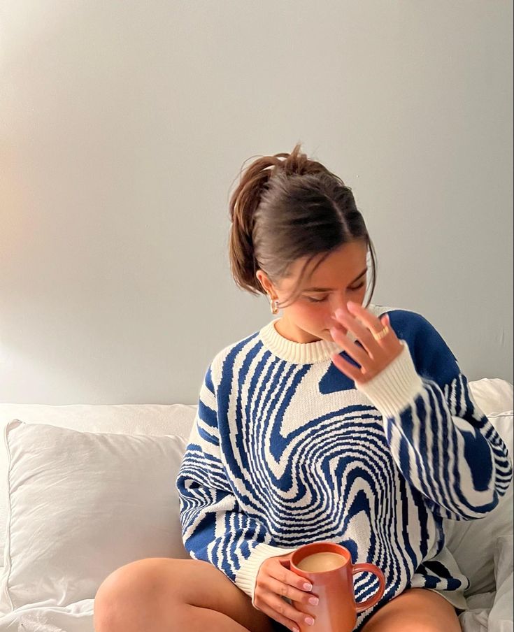 Woman wearing a soft blue and white swirl-patterned knitted pullover sweater while holding a mug on a bed.