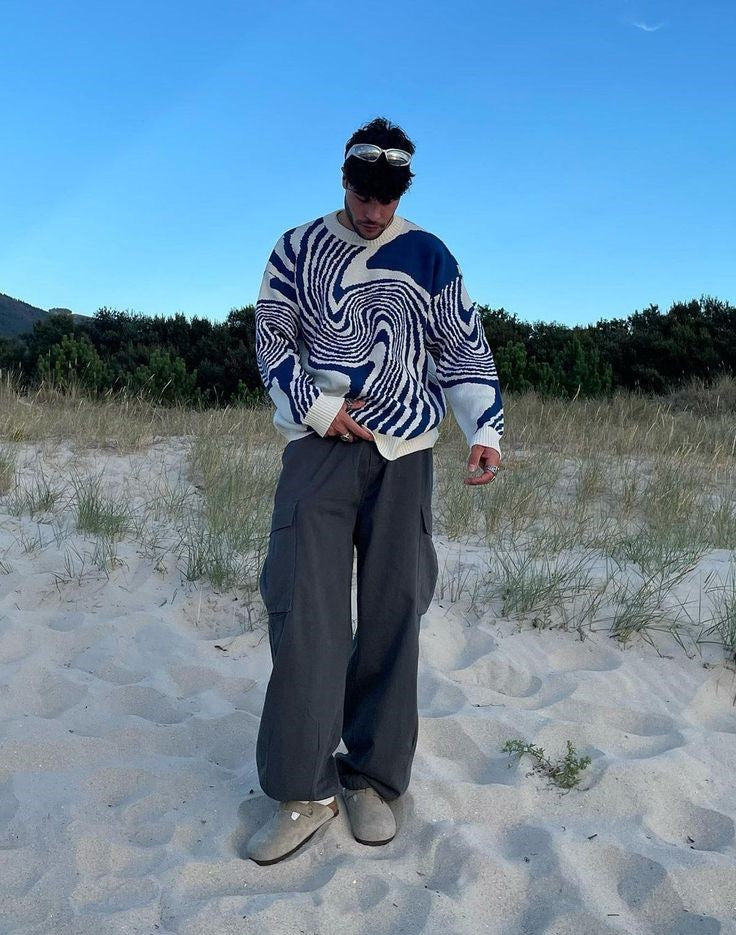 Person wearing a blue and white swirl-patterned knitted pullover sweater with grey cargo pants on a sandy beach.