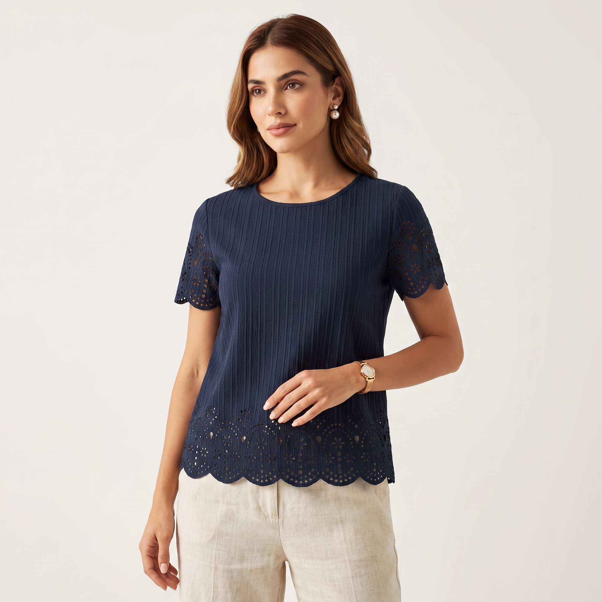 A woman modeling a navy blue short-sleeve top with scalloped eyelet lace trim at the hem and sleeves, paired with light beige pants, against a plain light gray studio background.