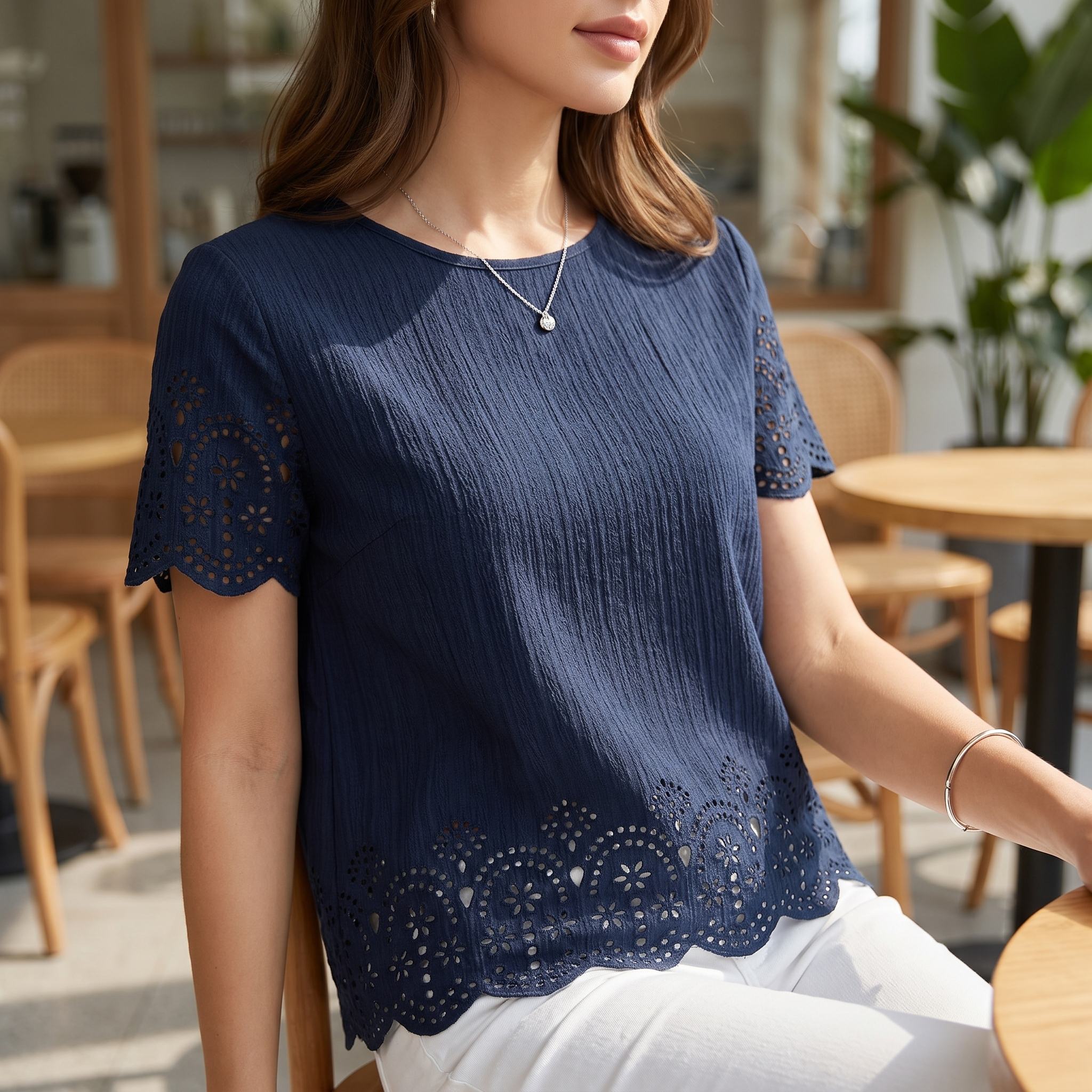 A woman wearing a navy blue short-sleeve top with eyelet lace detailing on the sleeves and hem, paired with white pants, seated in a cafe setting.