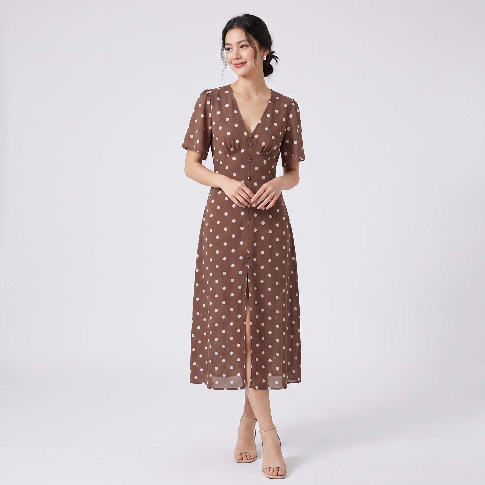A woman modeling a brown midi dress with a white polka dot pattern, short sleeves, and a V-neckline, standing against a light gray studio background.
