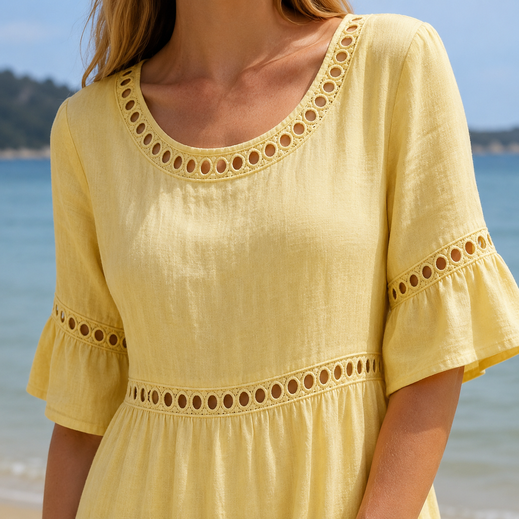 A woman wearing a pastel yellow linen-look dress with decorative eyelet trim around the scoop neckline, sleeves and empire waist, photographed in a sunny coastal setting. The close-up highlights the lightweight textured fabric, embroidered detailing and relaxed silhouette against a softly blurred seaside background.