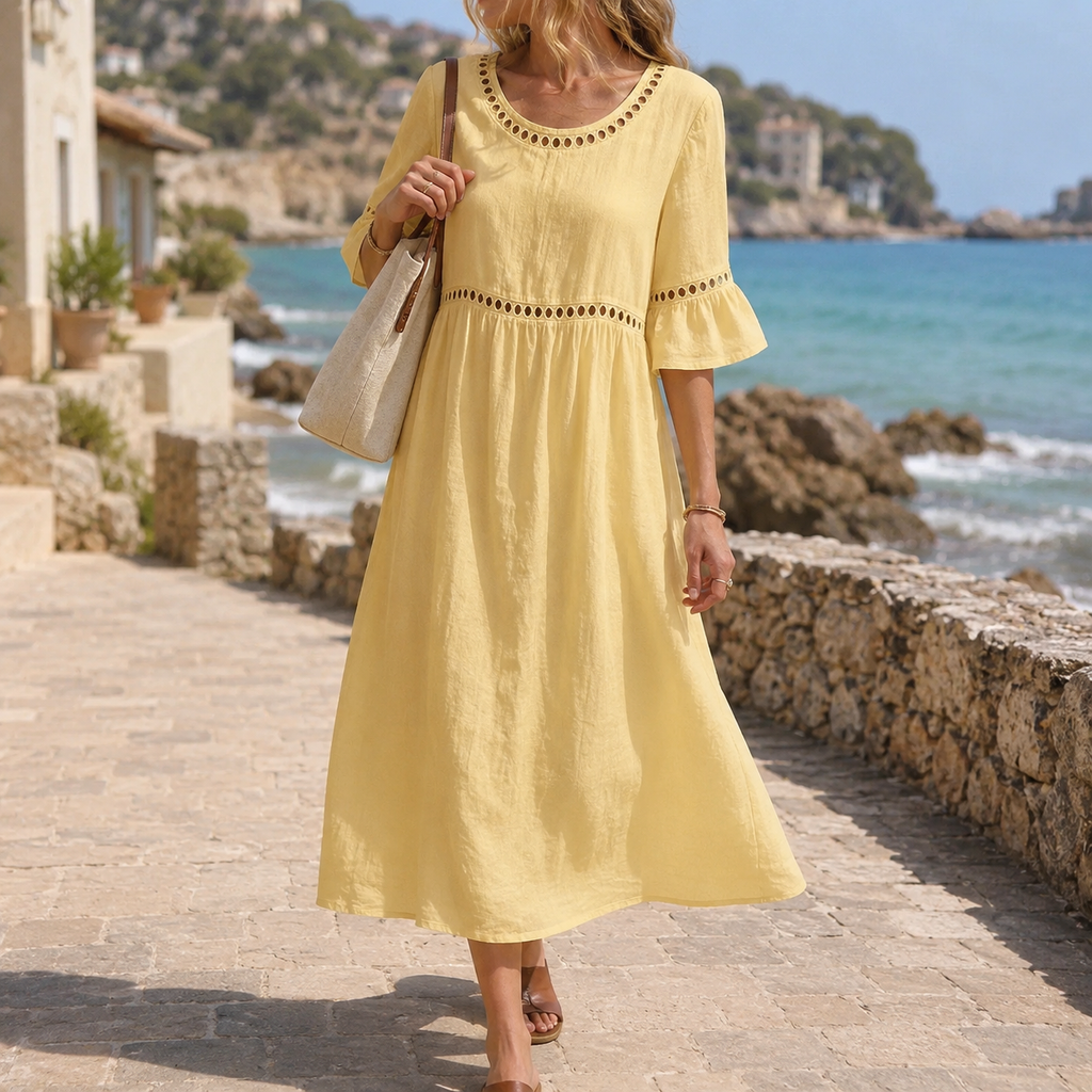 A woman wearing a pastel yellow linen-look midi dress with decorative eyelet trim around the scoop neckline, sleeves and empire waist walks along a sunlit coastal promenade. The flowing dress, textured fabric and relaxed silhouette are highlighted against a picturesque seaside town with turquoise water, rocky shoreline and stone buildings in the background.