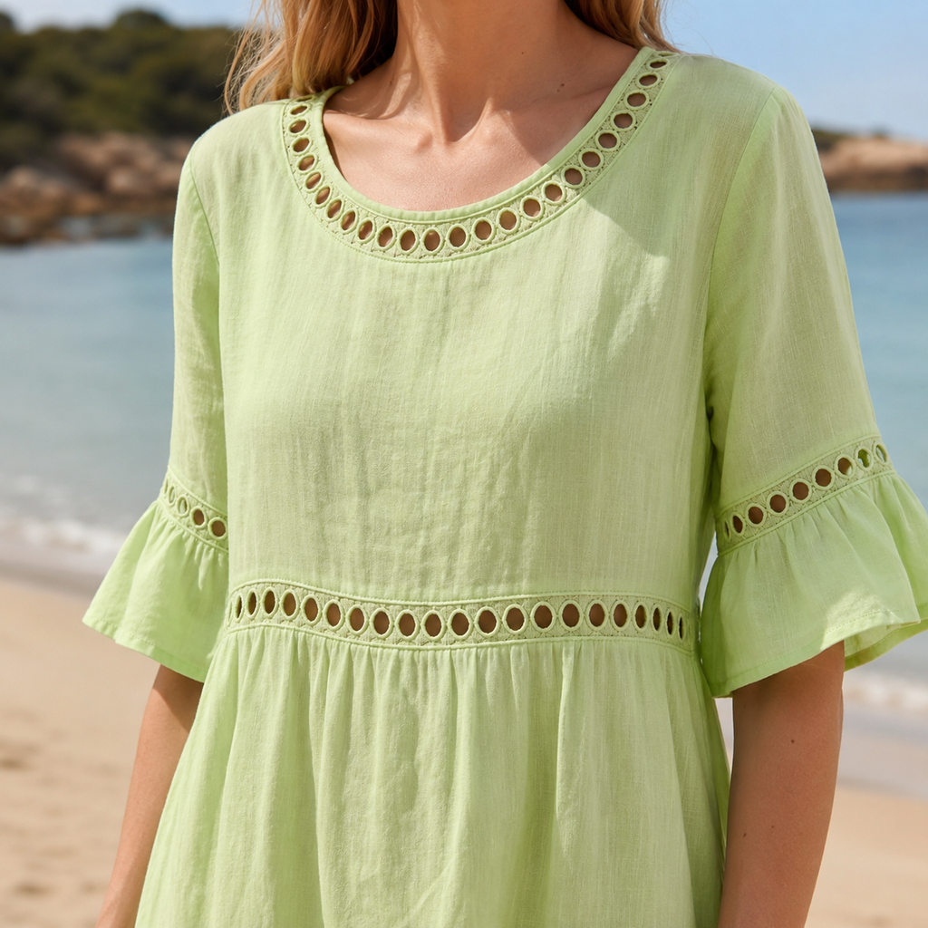 A woman wearing a light green linen-look dress with decorative eyelet trim around the scoop neckline, sleeves and empire waist stands on a sunny beach. The close-up highlights the lightweight textured fabric, embroidered detailing and relaxed silhouette against a softly blurred coastline and turquoise sea.