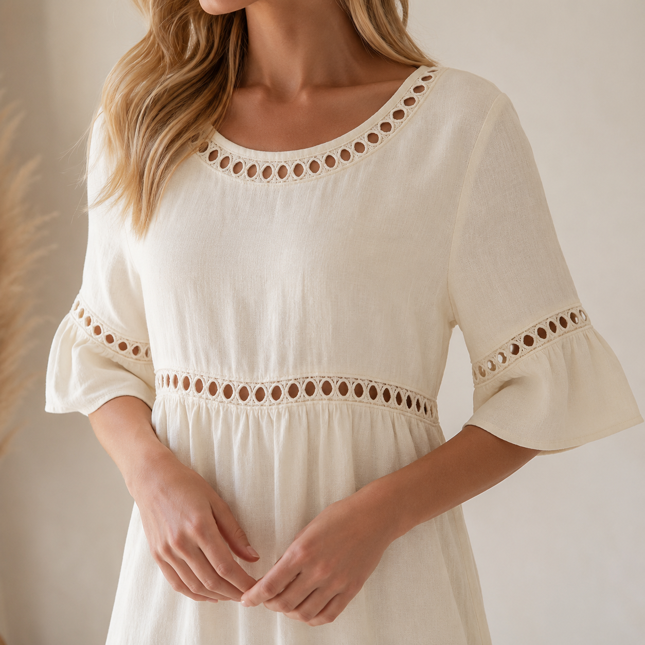 A woman wearing a cream-colored cotton dress with eyelet cutout details around the neckline, sleeves, and waist, set against a soft beige background.