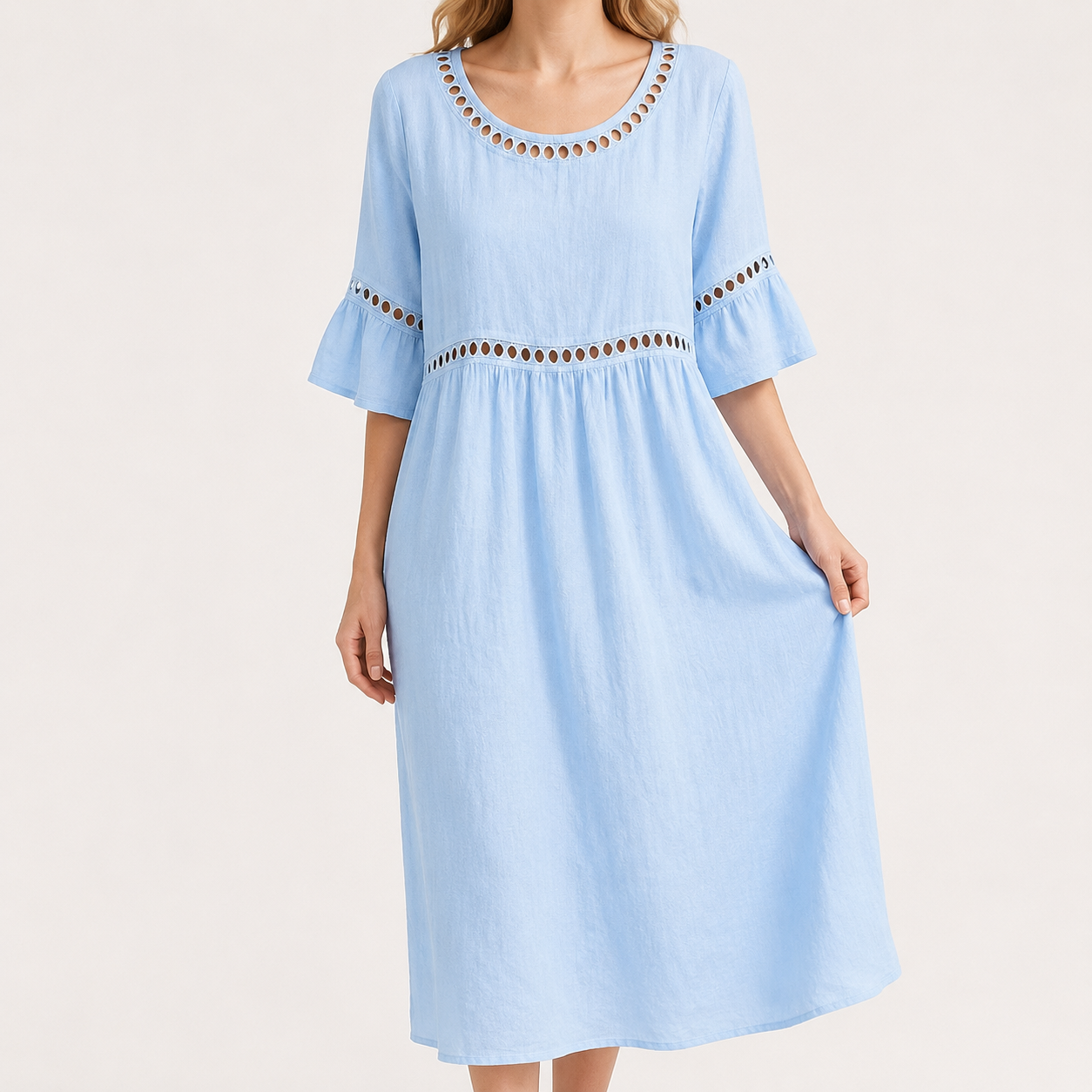 A woman wearing a light blue midi dress with three-quarter ruffled sleeves and decorative brown and gold cutout trim at the neckline, waist, and sleeve cuffs, photographed against a plain light beige studio background.