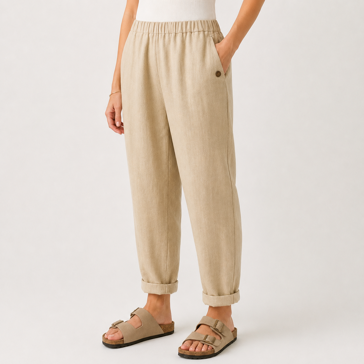 A person wearing loose beige linen pants with rolled cuffs and tan double-strap sandals, shown from the waist down against a clean off-white studio background.