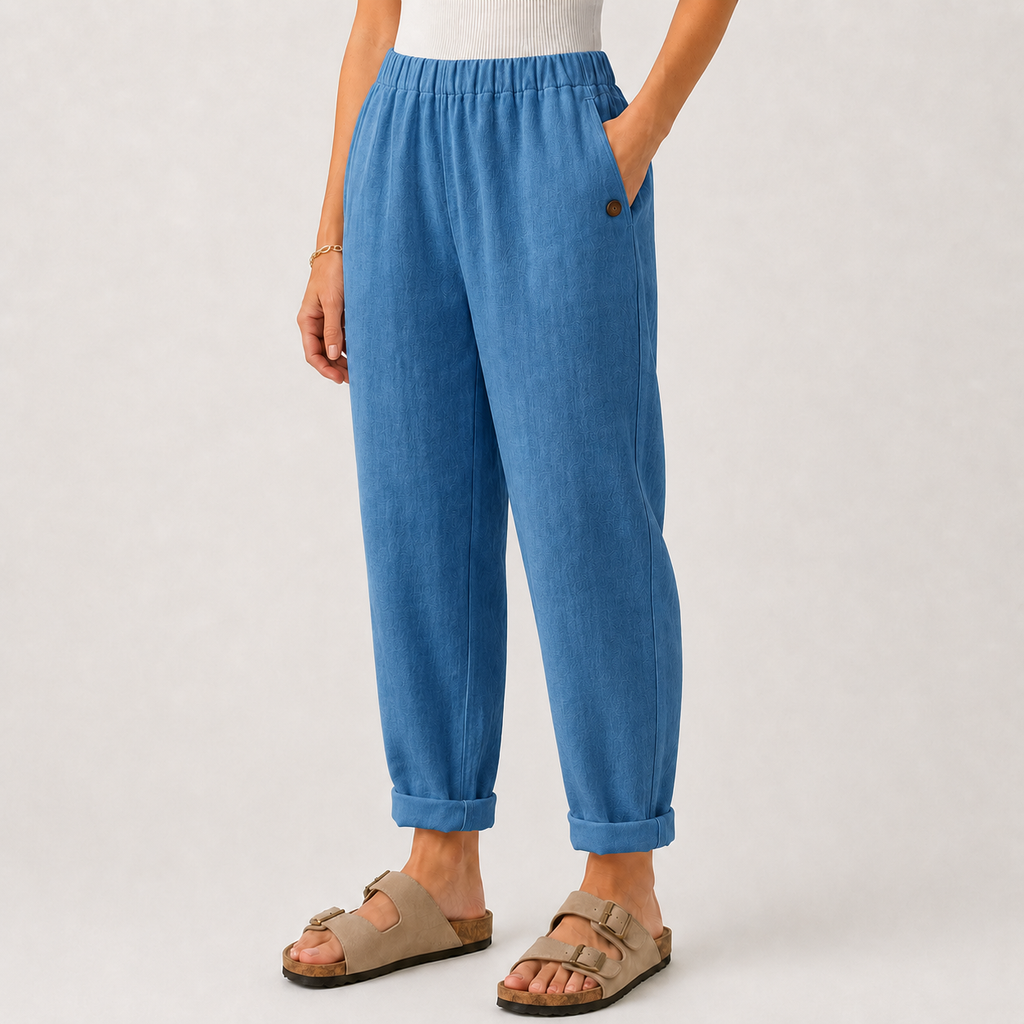 A person wearing blue relaxed-fit ankle-length pants with a cuffed hem, side pocket, and a small dark button detail, paired with beige double-strap sandals against a light gray studio background.