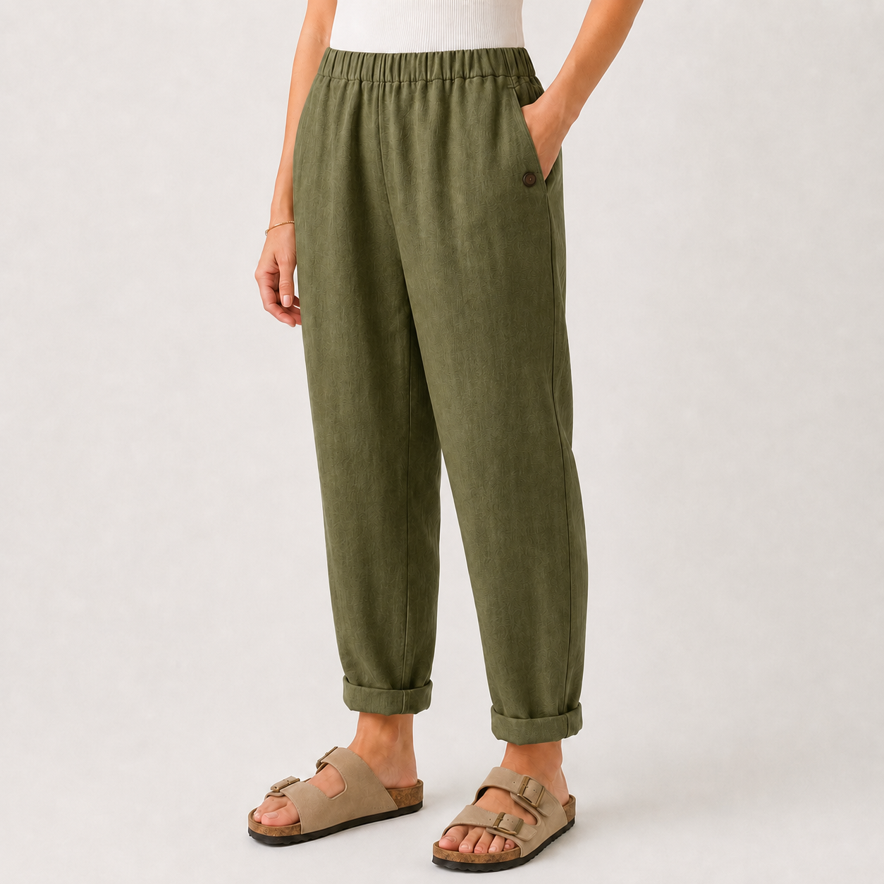 A person wearing olive green linen trousers with a relaxed, cuffed fit and side pockets, paired with beige double-strap sandals against a light gray studio background.