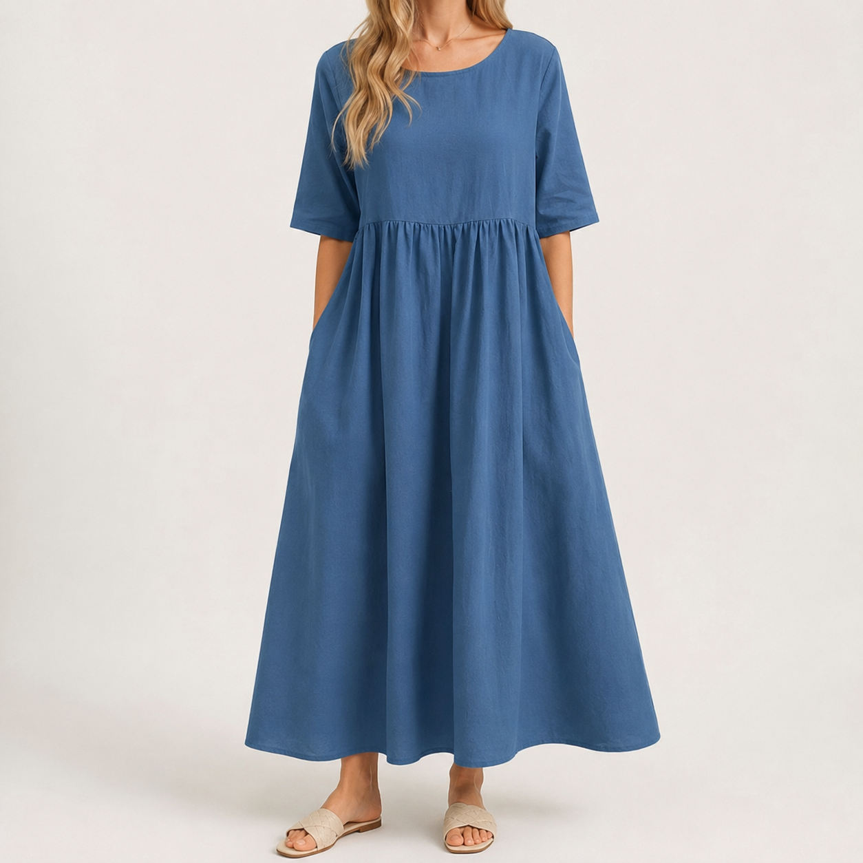 A woman wearing a dusty blue midi dress with short sleeves, a round neckline, and side pockets, paired with beige slide sandals, photographed against a plain off-white studio background.