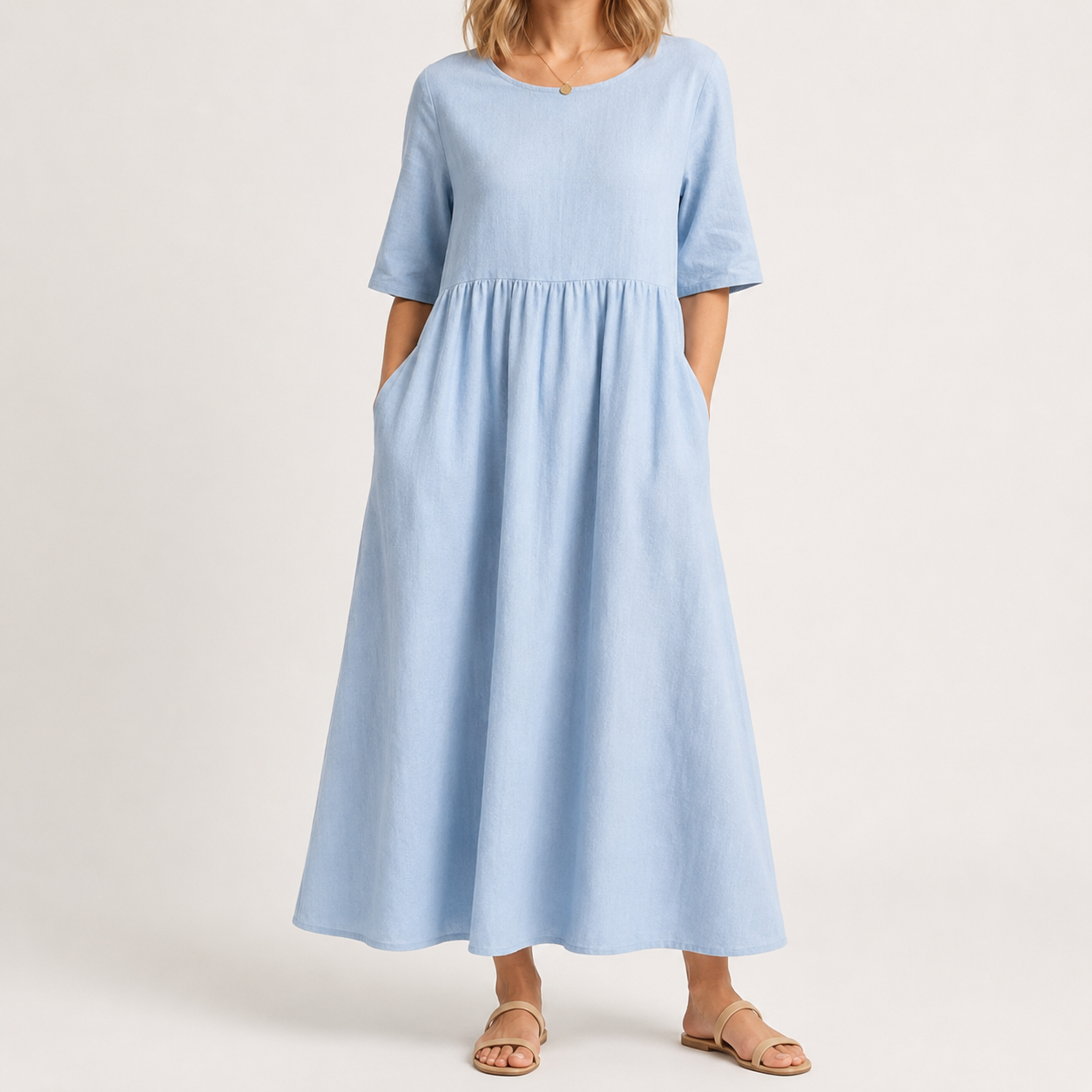 A woman wearing a light blue midi dress with short sleeves, a round neckline, and side pockets, paired with tan flat sandals, photographed against a plain light beige studio background.