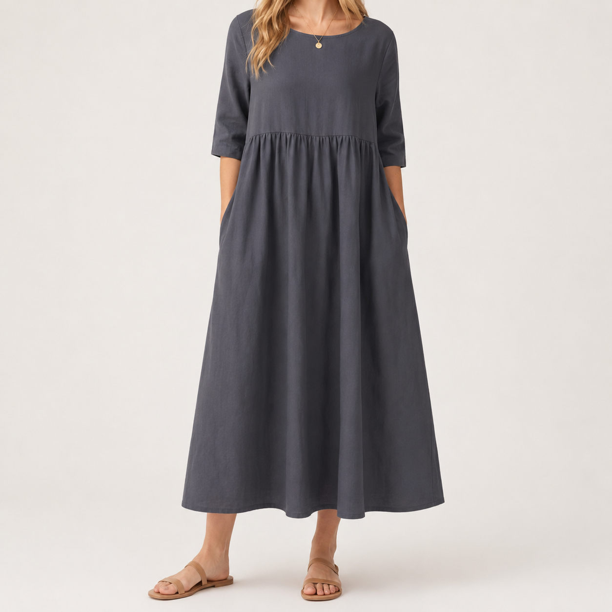 A woman wearing a dark gray midi dress with three-quarter sleeves, side pockets, and a gathered waist seam, paired with tan flat sandals against a light gray studio background.