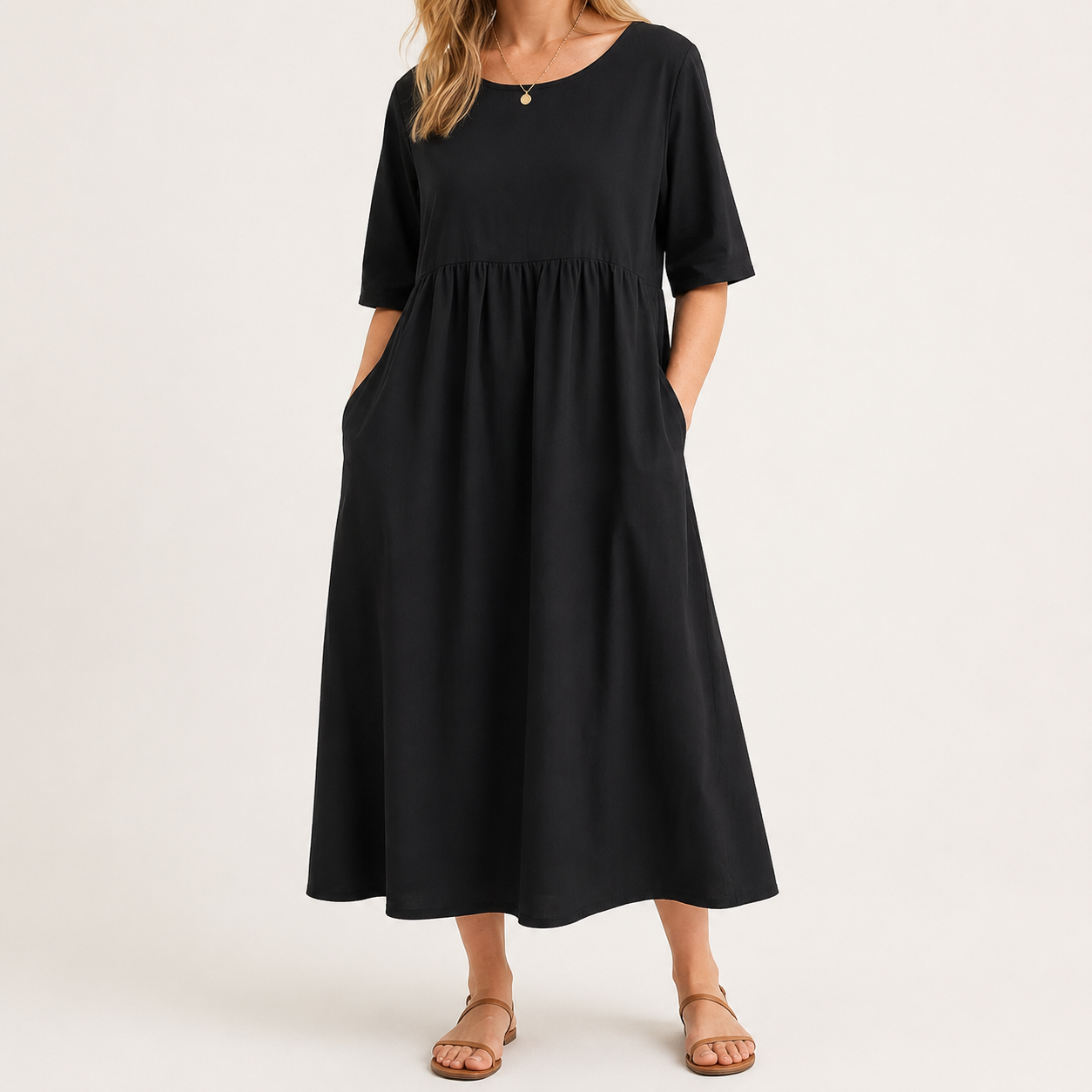 A woman wearing a black midi dress with short sleeves, a round neckline, side pockets, and a gathered waist, paired with tan flat sandals against a plain light background.