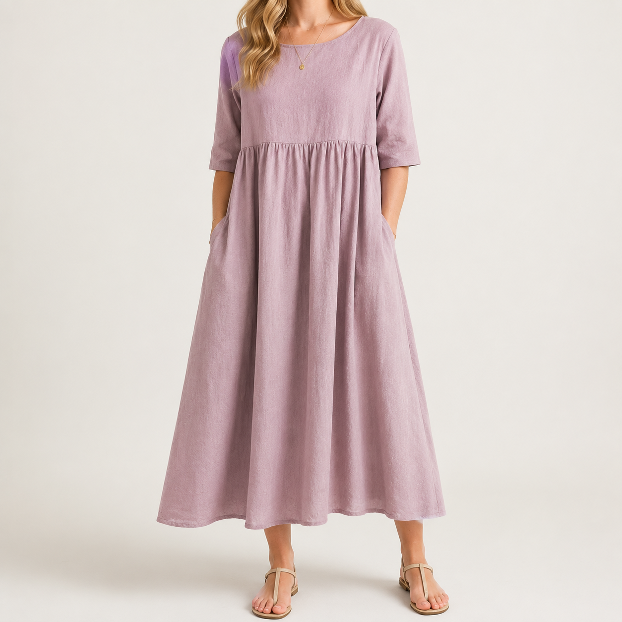 A woman wearing a dusty lavender midi dress with elbow-length sleeves, side pockets, and a gathered waist, paired with neutral sandals against a light beige studio background.