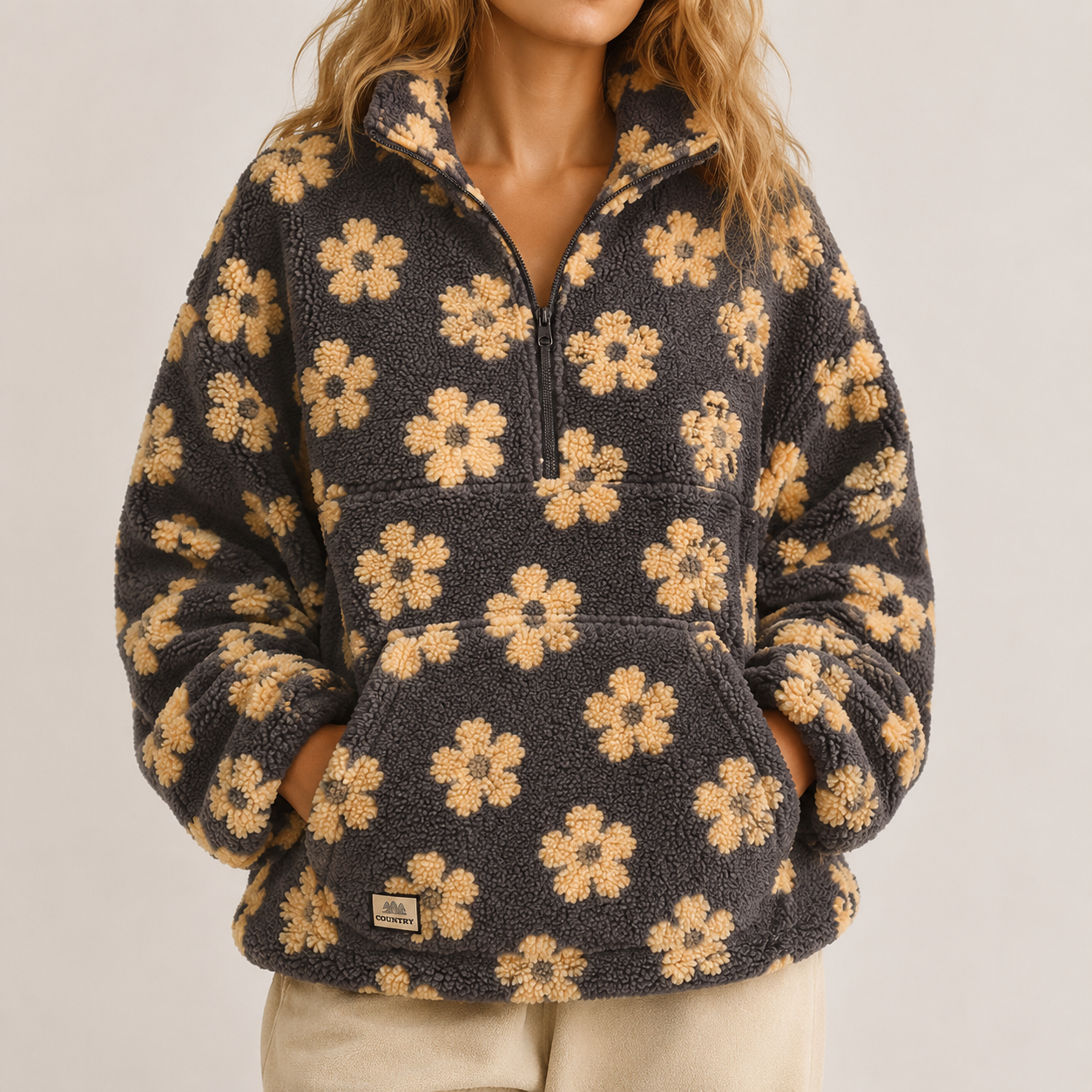 A woman wearing a dark charcoal fleece quarter-zip pullover with a tan floral pattern, paired with light beige pants, against a plain light gray background.