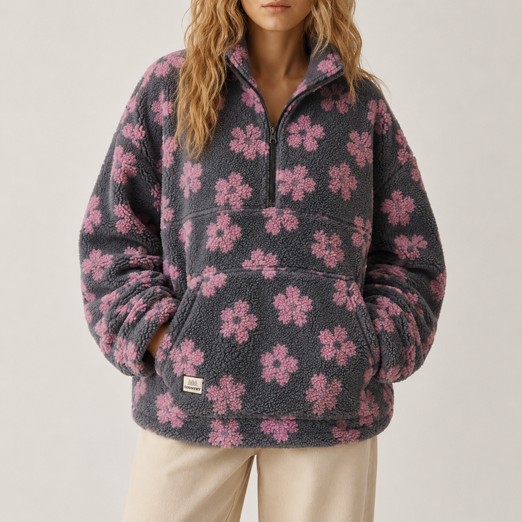A woman wearing a dark gray fleece pullover with a pink floral pattern, paired with cream pants, against a plain light gray studio background.