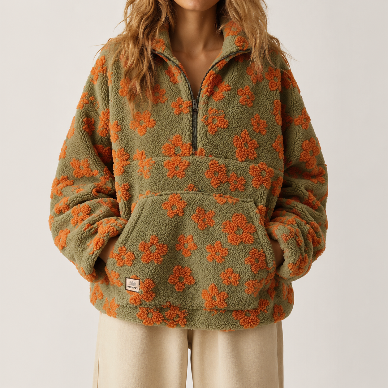 A person wearing a fuzzy quarter-zip fleece pullover in olive green with an orange floral pattern, paired with light beige pants, against a plain light gray studio background.