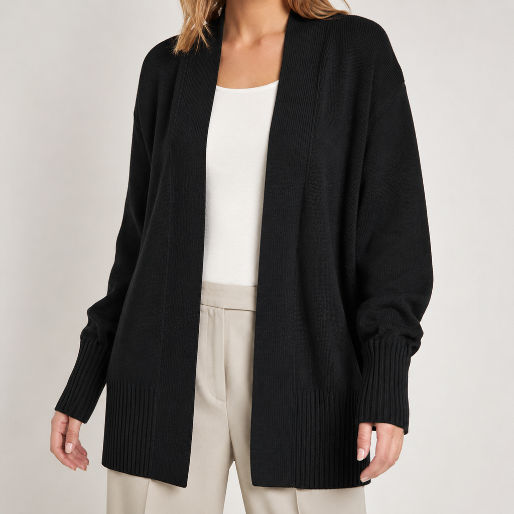 A woman wearing a black open-front knit cardigan over a white top and light beige trousers, photographed against a plain light gray studio background.