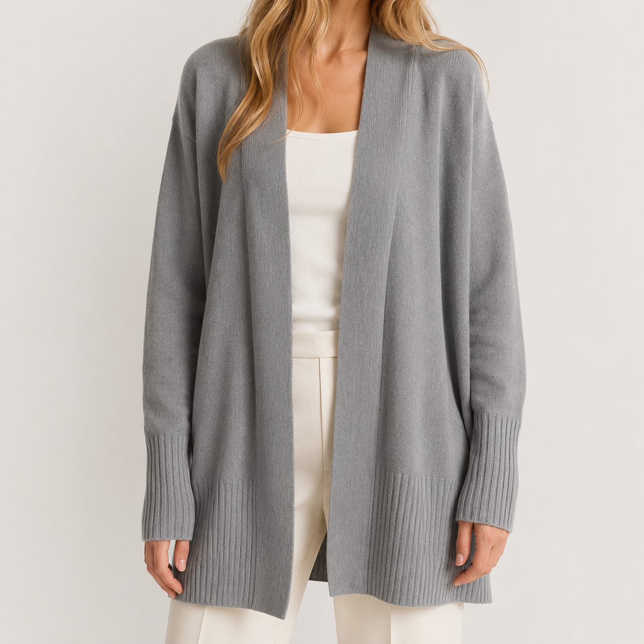 A woman wearing a light gray open-front knit cardigan over a white top and white pants, photographed against a plain light gray studio background.