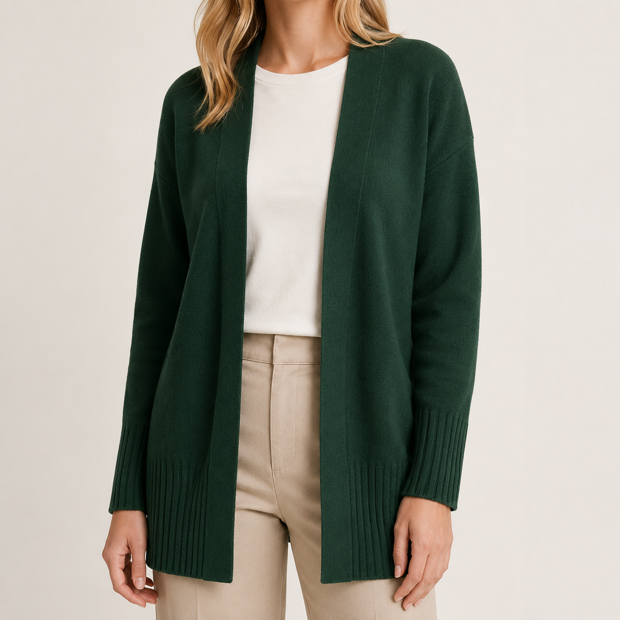 A woman wearing a dark forest green open-front knit cardigan over a white top and beige trousers, photographed against a plain light background.