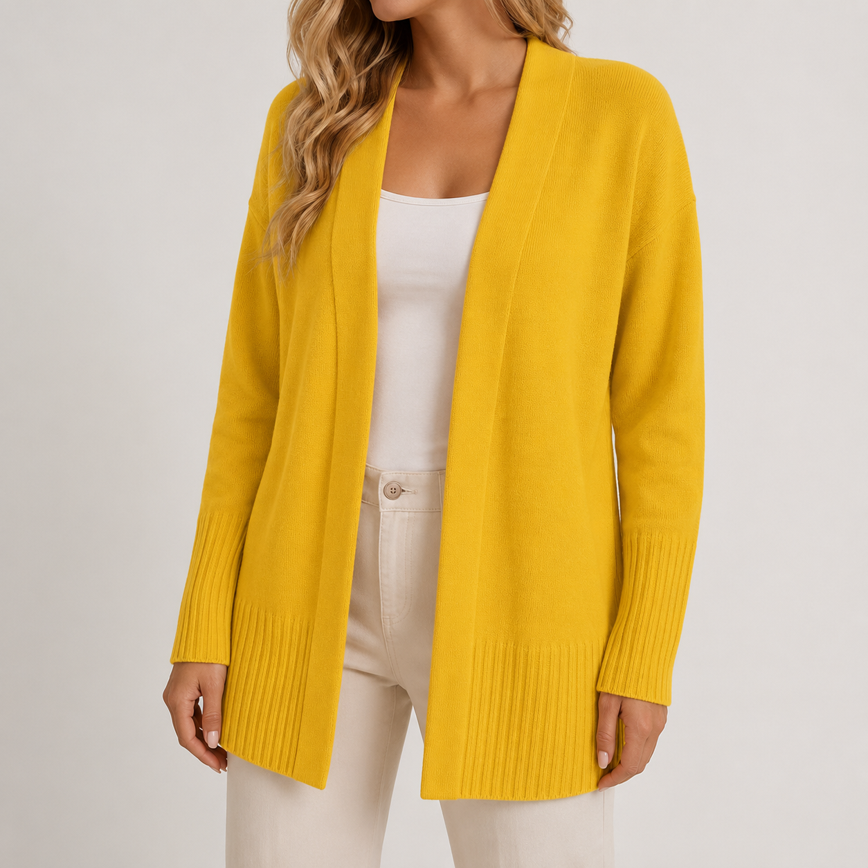 A woman wearing a bright yellow open-front knit cardigan over a white top and cream pants, photographed against a plain light gray studio background.