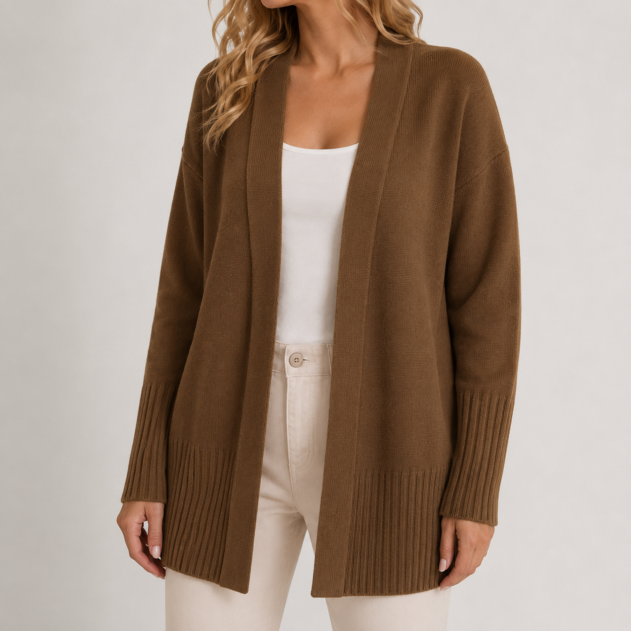 A woman wearing a brown open-front knit cardigan over a white tank top and cream pants, photographed against a plain light gray studio background.