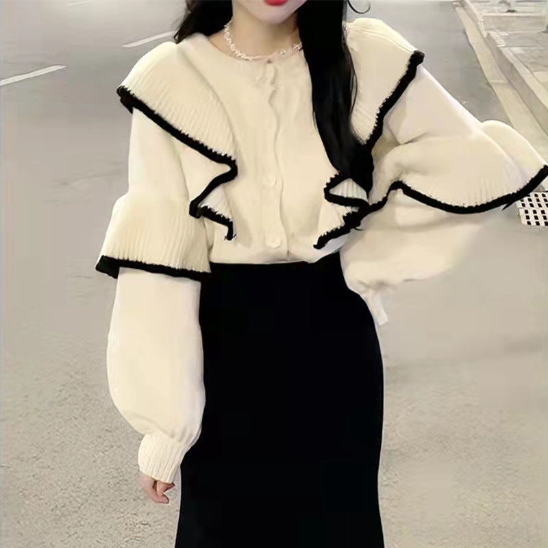 Cream knit sweater with black-trimmed ruffled collar and sleeves, paired with a black skirt for an elegant look.