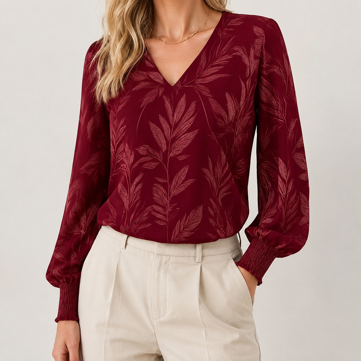 A woman wearing a long-sleeve burgundy blouse with a tonal leaf pattern and cream trousers, shown from the shoulders to the hips against a light gray studio background.