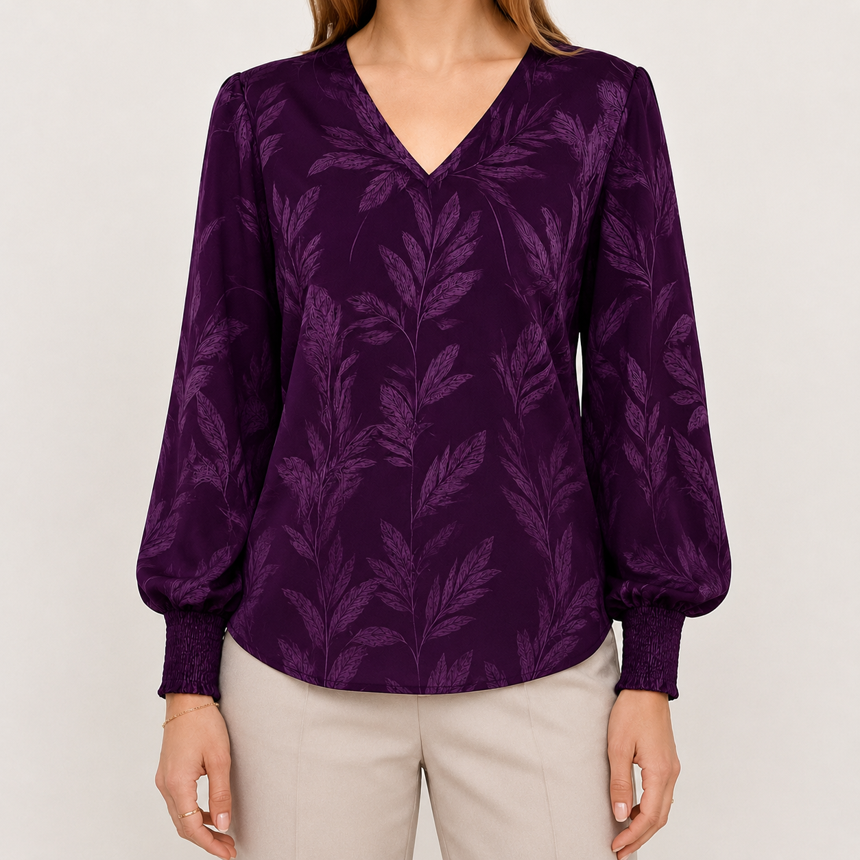 A woman wearing a long-sleeve V-neck blouse in deep purple with a tonal leaf pattern, paired with light beige pants, against a plain light gray background.