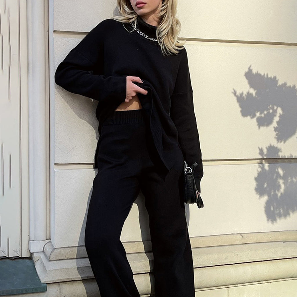 Black relaxed-fit turtleneck sweater with side splits, worn with matching knit trousers and a silver chain necklace.